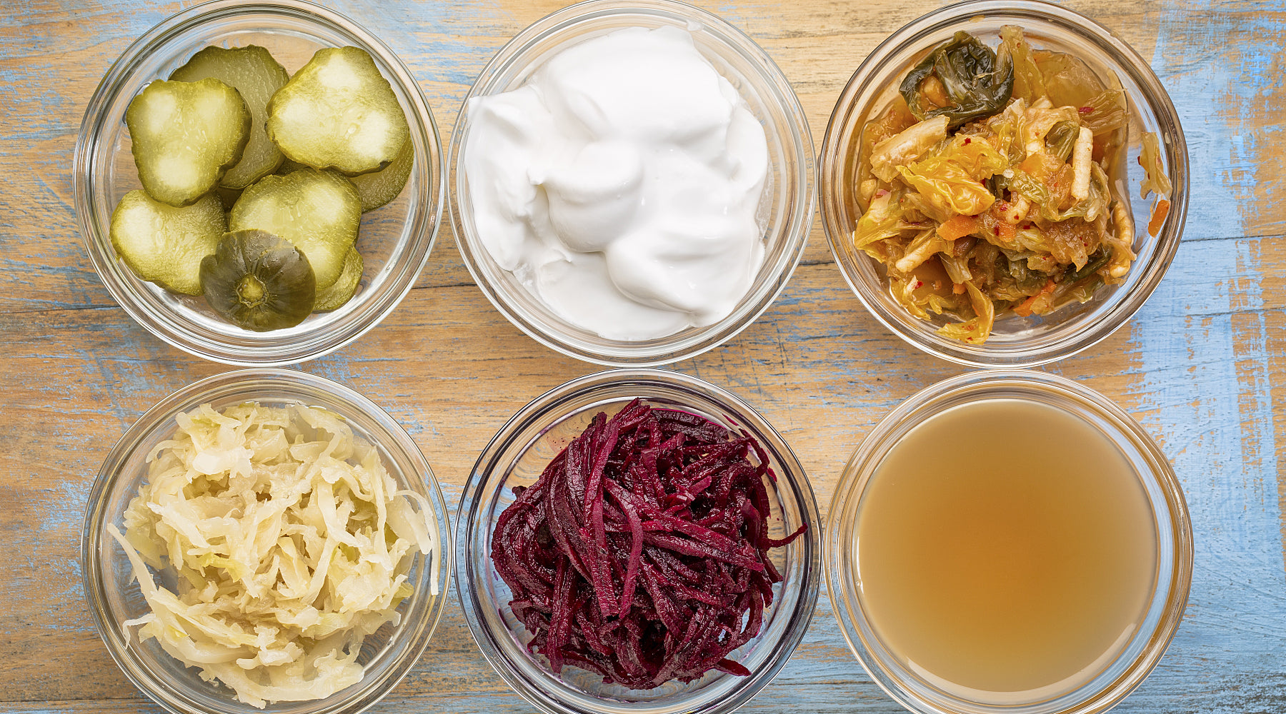 Six glass bowls of fermented foods, including pickles, yogurt, kimchi and beets