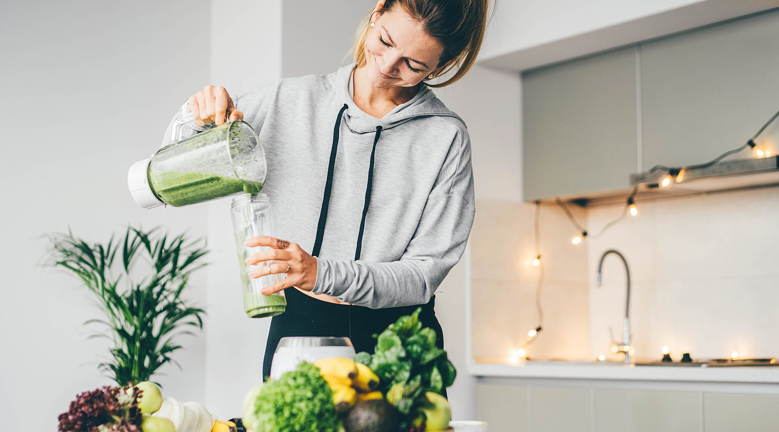 Young woman making a smoothie using a blender with fruits and vegetables on her kitchen countertop.