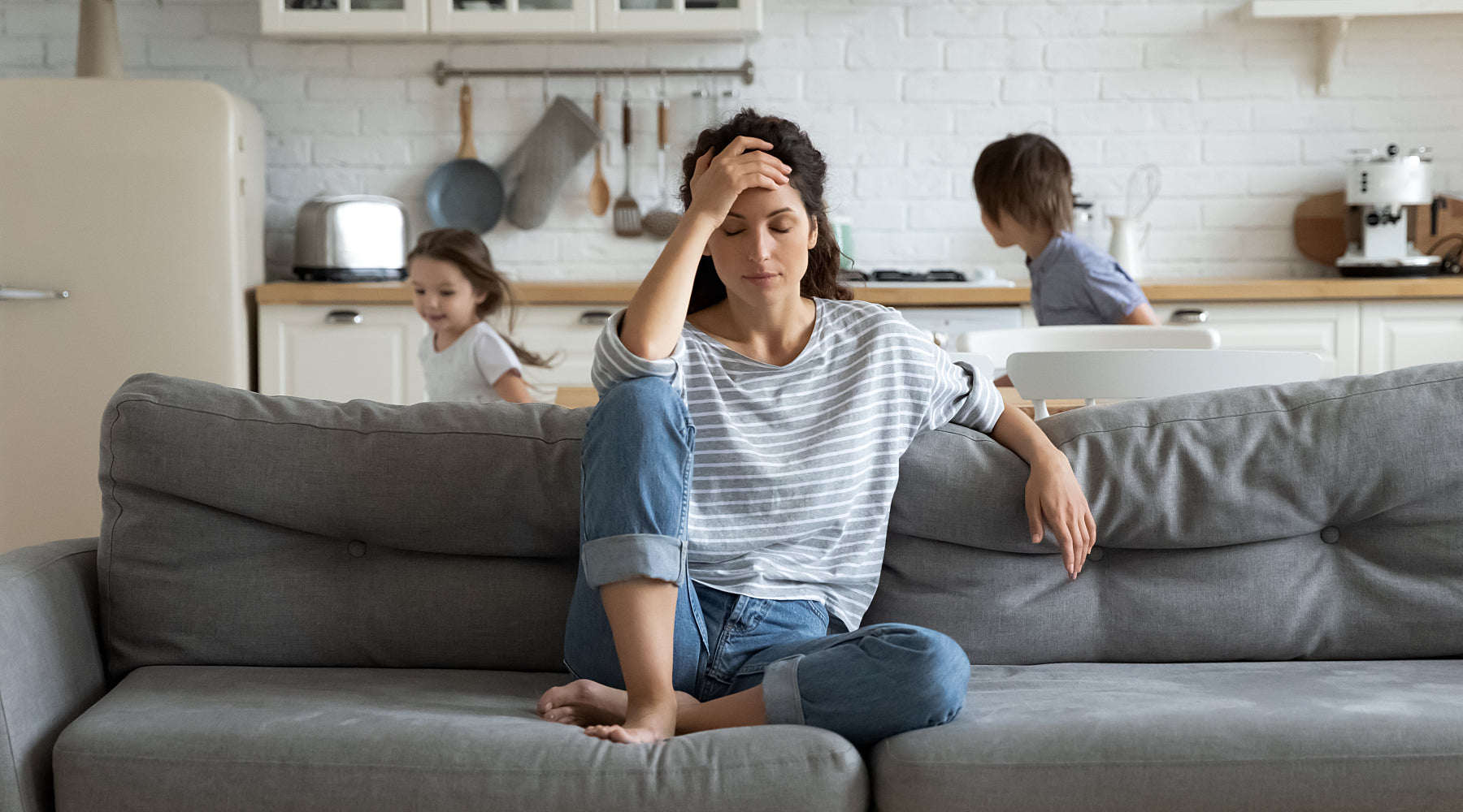 Young mother sitting on a couch looking stressed with two kids in the background.